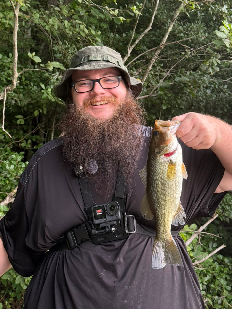 The Owner Of FusedOutdoors wearing a camouflage bucket hat, glasses, and a black shirt holds up a freshly caught Largemouth Bass fish outdoors in front of a dense green forest background.