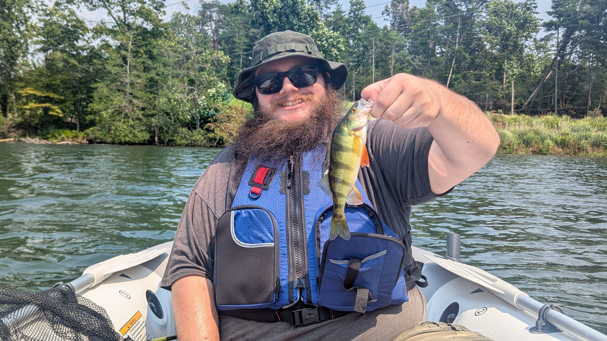 The Owner of FusedOutdoors wearing a bucket hat, sunglasses, and a blue life vest sitting in a boat on the water, holding up a yellow Perchthat he just caught. Trees line the riverbank in the background.